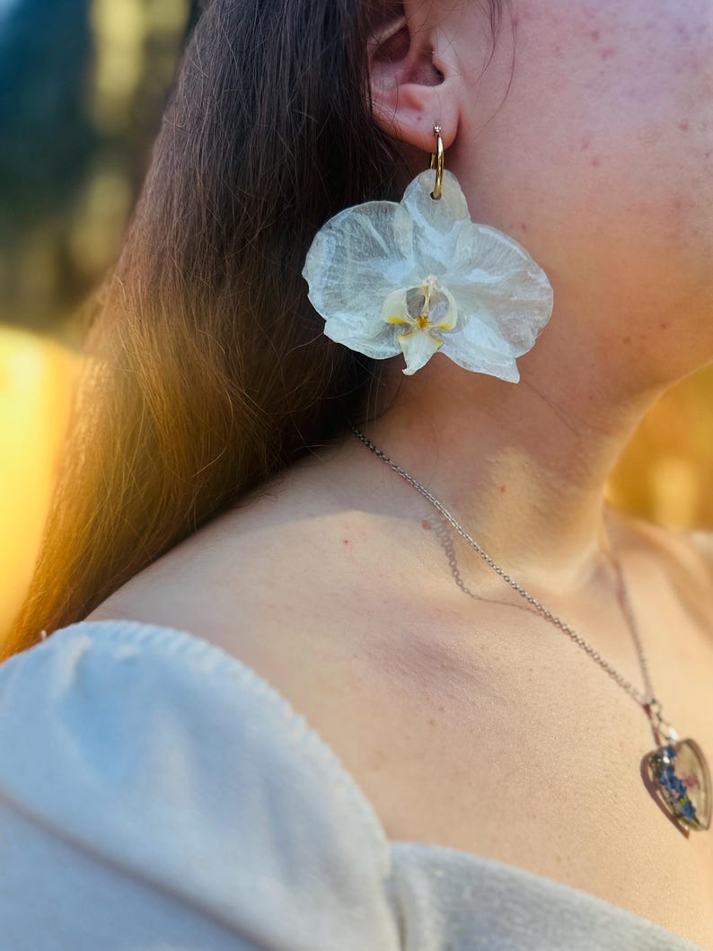 Pode incluir: Um brinco grande em forma de flor de orqu&iacute;dea branca transl&uacute;cida com um centro amarelo e roxo, suspenso por um aro fino de tom dourado. O brinco apresenta texturas de p&eacute;talas delicadas e realistas, exibindo um design bot&acirc;nico.