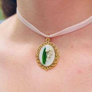 Pode incluir: Um colar com um pingente oval dourado em uma fita branca. O pingente apresenta um fundo branco com uma folha verde e flores brancas de l&iacute;rio do vale.