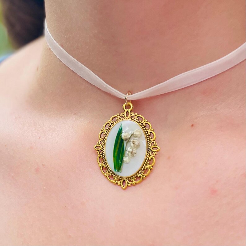 Pode incluir: Um colar com um pingente oval dourado em uma fita branca. O pingente apresenta um fundo branco com uma folha verde e flores brancas de l&iacute;rio do vale.