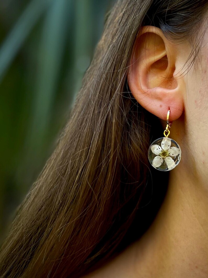Puede incluir: Un par de pendientes de aro dorados con una flor blanca prensada dentro de un c&iacute;rculo de resina transparente.