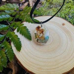 May include: A delicate necklace with a resin pendant featuring dried flowers and mushrooms. The pendant is oval-shaped and has a clear resin finish, showcasing the natural elements inside.