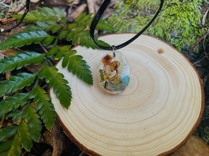 May include: A delicate necklace with a resin pendant featuring dried flowers and mushrooms. The pendant is oval-shaped and has a clear resin finish, showcasing the natural elements inside.