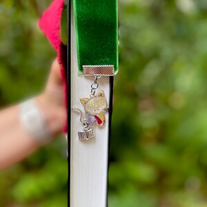 May include: A green velvet bookmark with a silver charm of a white cat holding a book. The cat charm has pressed flowers inside.