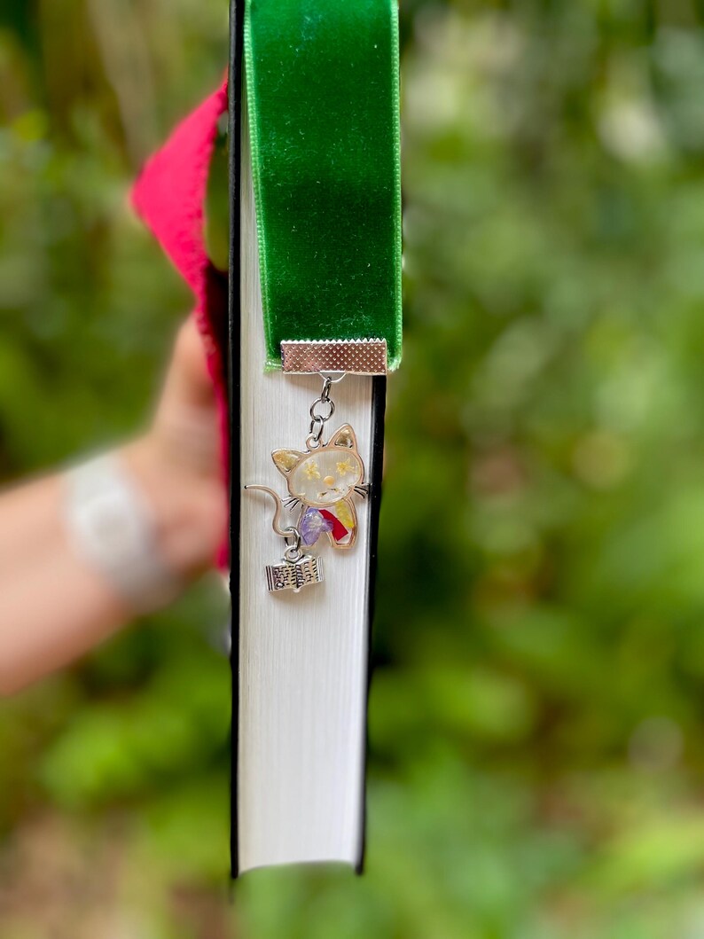 May include: A green velvet bookmark with a silver charm of a white cat holding a book. The cat charm has pressed flowers inside.