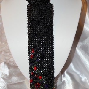 May include: A black beaded necklace with a tie-like design, displayed on a white mannequin. The lower portion of the necklace is adorned with red, green, and purple accents, creating a floral pattern. The necklace is made of small, round beads.