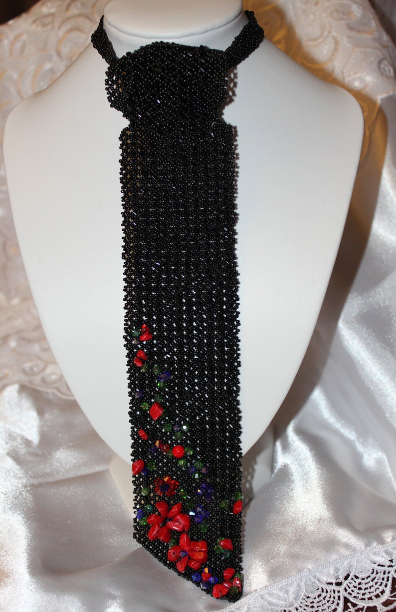 May include: A black beaded necklace with a tie-like design, displayed on a white mannequin. The lower portion of the necklace is adorned with red, green, and purple accents, creating a floral pattern. The necklace is made of small, round beads.