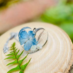 Könnte beinhalten: Eine herzförmige Halskette mit einem Anhänger, in dem eine blaue Blume und weiße Blumen in klarem Harz gepresst sind. Der Anhänger ist an einer silbernen Kette.