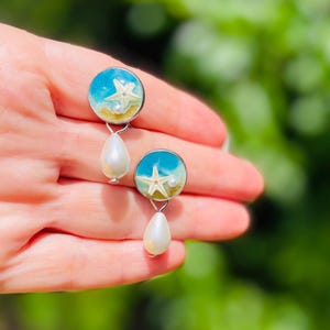 Op de afbeelding: Een paar oorbellen met een strandthema. Elke oorbel heeft een ronde, blauwe hars top met een zeester- en zandontwerp. Onder elke ronde top hangt een witte, druppelvormige parel. De oorbellen worden in een hand gehouden tegen een vage groene achtergrond.