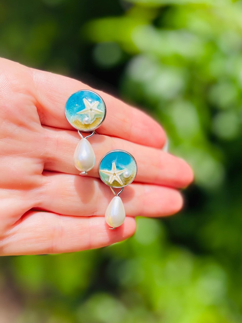 Op de afbeelding: Een paar oorbellen met een strandthema. Elke oorbel heeft een ronde, blauwe hars top met een zeester- en zandontwerp. Onder elke ronde top hangt een witte, druppelvormige parel. De oorbellen worden in een hand gehouden tegen een vage groene achtergrond.