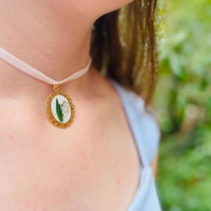 Pode incluir: Um colar de choker de cetim branco com um pingente oval dourado que apresenta uma flor de l&iacute;rio do vale prensada.