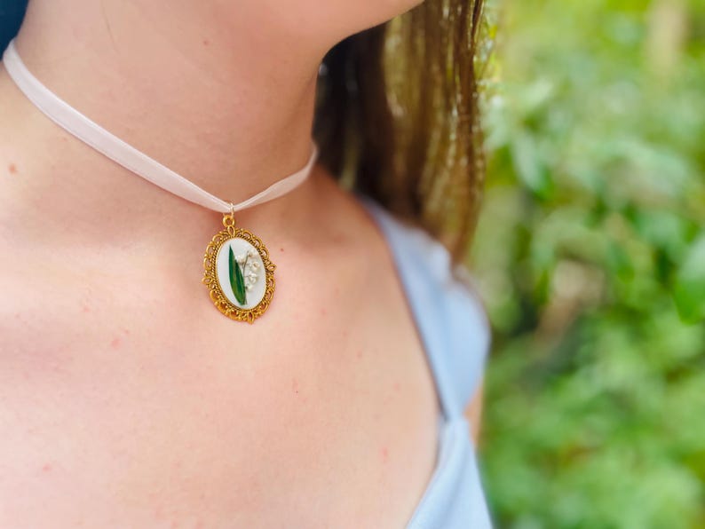 Pode incluir: Um colar de choker de cetim branco com um pingente oval dourado que apresenta uma flor de l&iacute;rio do vale prensada.