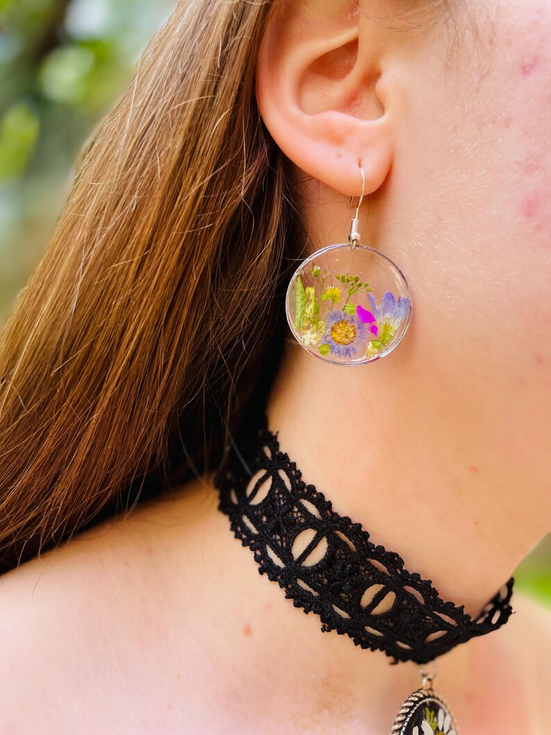 Puede incluir: Pendientes circulares transparentes con flores secas en tonos morados, amarillos y verdes. Los pendientes cuelgan de ganchos plateados. Se ve un collar de gargantilla de encaje negro.
