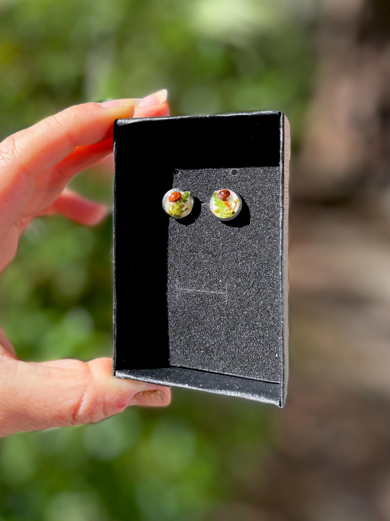 May include: A pair of small, round earrings with a clear resin base and a small, colorful flower design inside. The earrings are displayed in a black box.