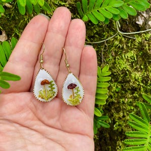 Puede incluir: Un par de pendientes en forma de l&aacute;grima con ganchos de metal dorado. Cada pendiente presenta un fondo blanco con un peque&ntilde;o hongo rojo y marr&oacute;n y follaje verde prensado en su interior.