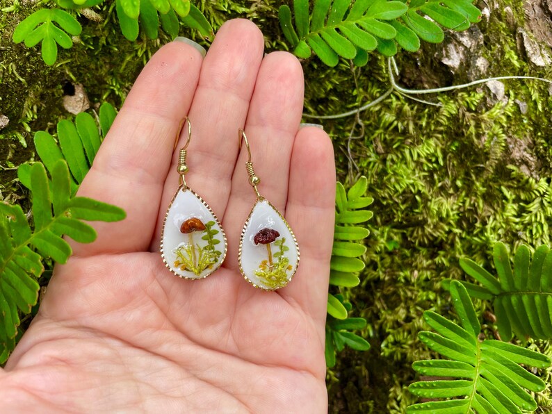 Puede incluir: Un par de pendientes en forma de l&aacute;grima con ganchos de metal dorado. Cada pendiente presenta un fondo blanco con un peque&ntilde;o hongo rojo y marr&oacute;n y follaje verde prensado en su interior.