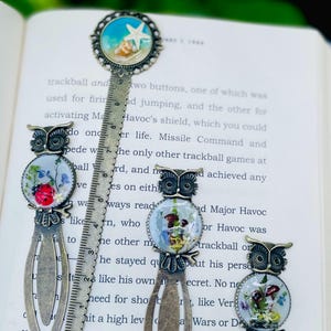 May include: A collection of decorative metal bookmarks with owl and beach-themed designs. The bookmarks feature intricate details and are set against the backdrop of an open book.