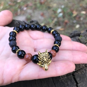 May include: A black and brown beaded bracelet with a gold wolf charm. The bracelet is made with black lava rock beads and brown sandstone beads.