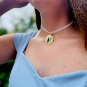 Pode incluir: Um close-up de uma mulher vestindo um vestido azul claro com um colar branco de choker e um pingente dourado com um design verde e branco.