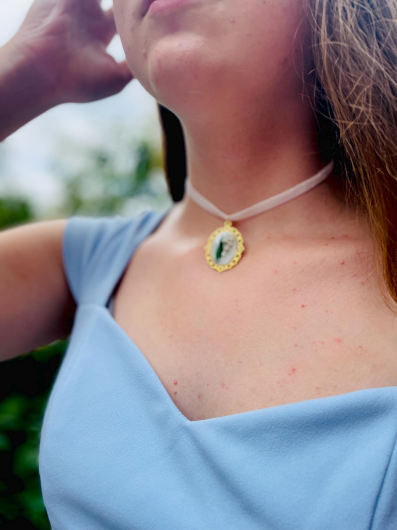 Pode incluir: Um close-up de uma mulher vestindo um vestido azul claro com um colar branco de choker e um pingente dourado com um design verde e branco.