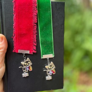 May include: Two velvet ribbon bookmarks with a black cat charm. The bookmarks are red and green. The charms have a silver finish and feature a black cat with white stars.