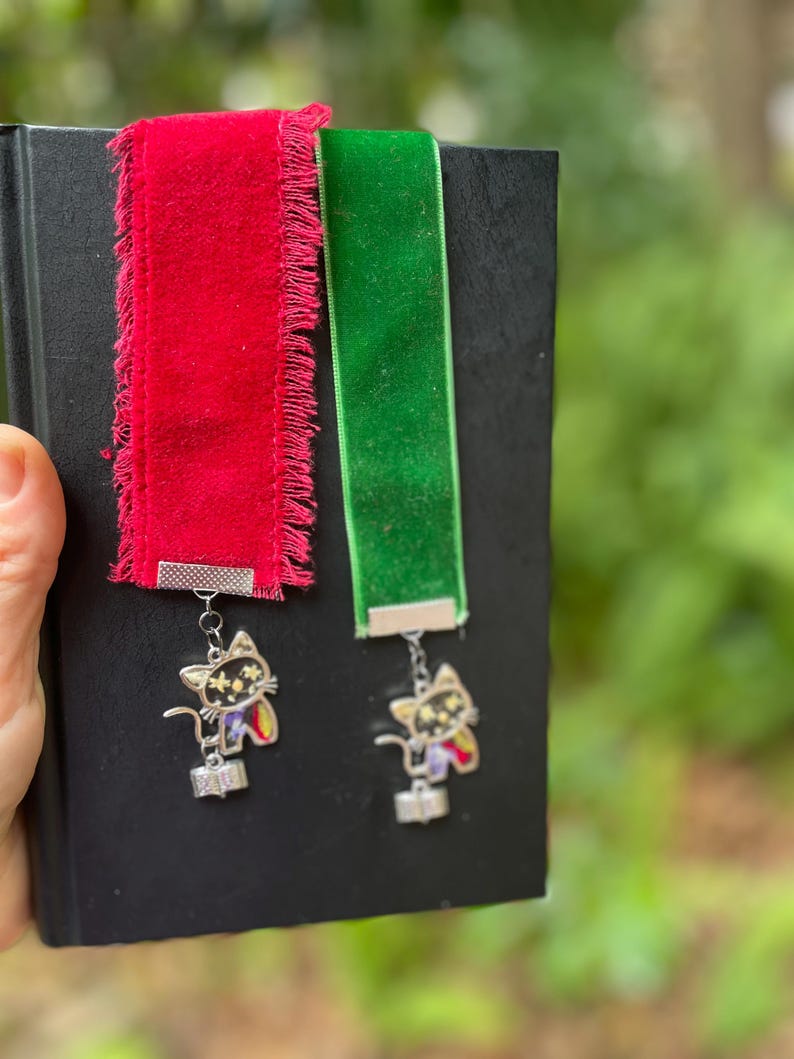 May include: Two velvet ribbon bookmarks with a black cat charm. The bookmarks are red and green. The charms have a silver finish and feature a black cat with white stars.