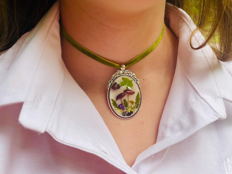 K&ouml;nnte beinhalten: Eine gr&uuml;ne Samt-Chokerkette mit einem silbernen ovalen Anh&auml;nger mit einem gepressten Blumen- und Pilzdesign.