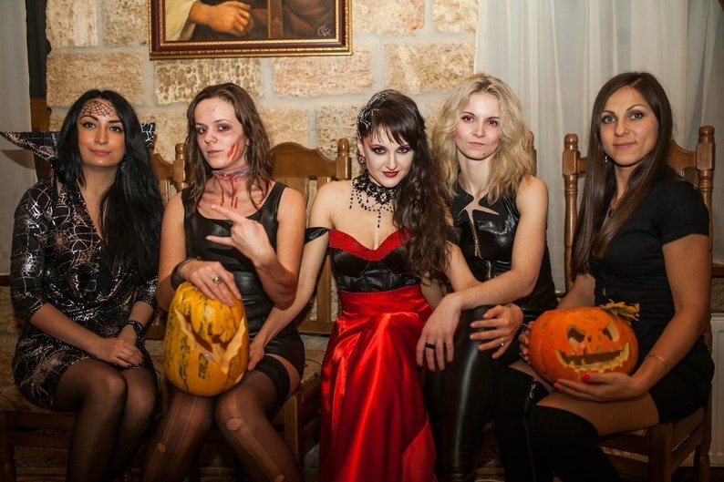 May include: Five women dressed in Halloween costumes, sitting on chairs in front of a brick wall. They are holding carved pumpkins. One woman is dressed as a vampire, another as a zombie, and the others are in black dresses.