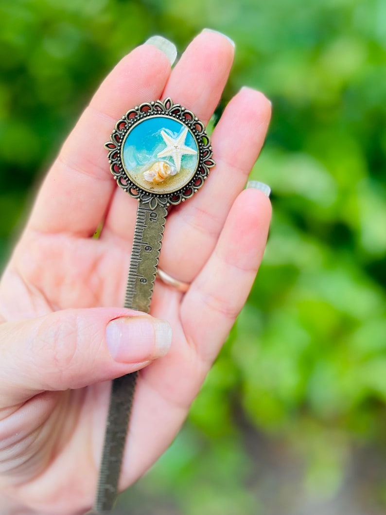 May include: A bronze metal bookmark with a decorative circular top featuring a miniature beach scene. The scene includes a white starfish, seashells, and blue water. The bookmark has a ruler design along its length.