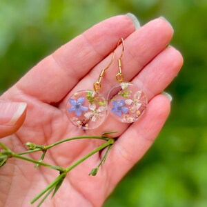 Puede incluir: Un par de pendientes de aro dorados con colgantes redondos de resina transparente. Cada colgante presenta flores azules y blancas prensadas.