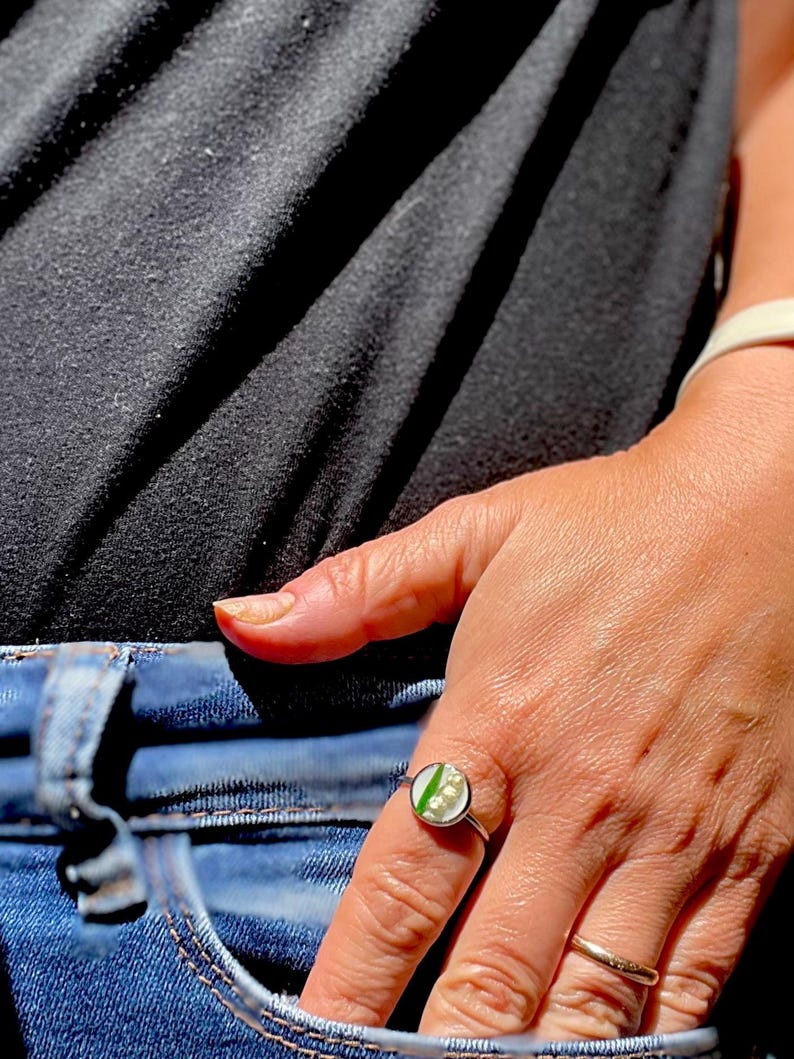 May include: A silver ring with a white and green resin inlay featuring a floral design. The ring is worn on a person's finger.