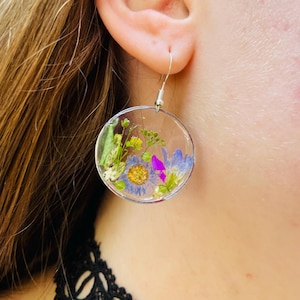 Puede incluir: Pendientes circulares transparentes con flores secas encapsuladas en resina. Los pendientes presentan una variedad de flores coloridas, incluyendo flores azules, moradas y amarillas, con ganchos plateados. Un accesorio &uacute;nico y hecho a mano.