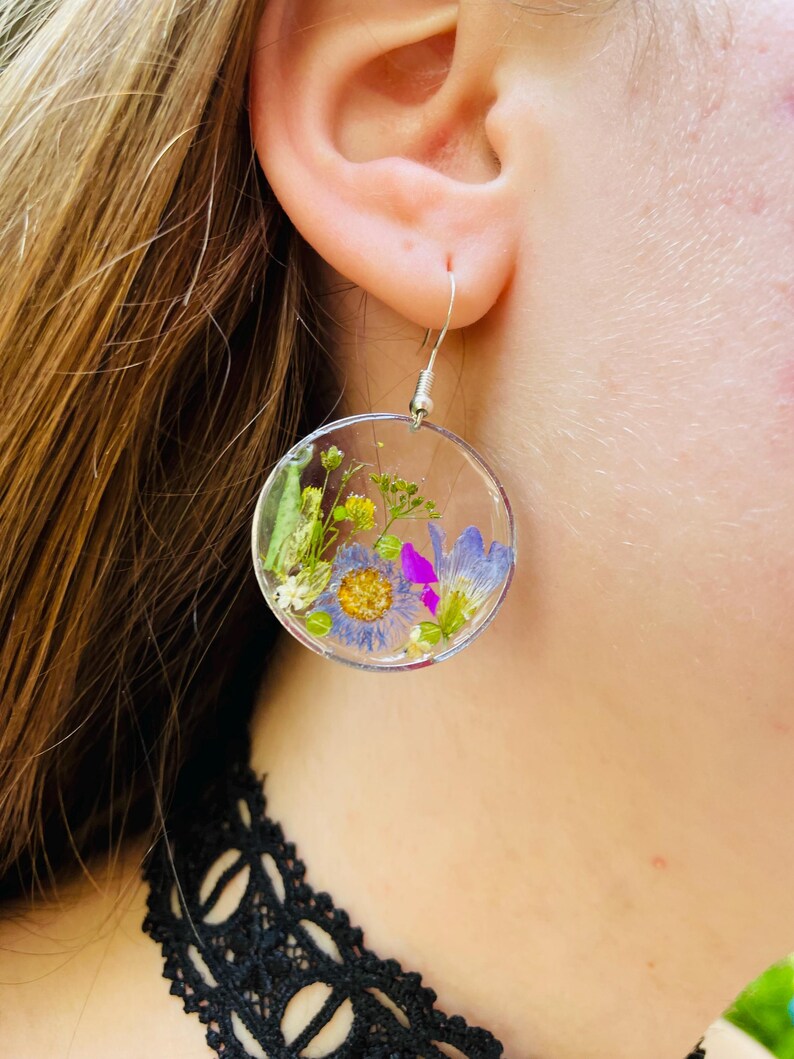 Puede incluir: Pendientes circulares transparentes con flores secas encapsuladas en resina. Los pendientes presentan una variedad de flores coloridas, incluyendo flores azules, moradas y amarillas, con ganchos plateados. Un accesorio &uacute;nico y hecho a mano.