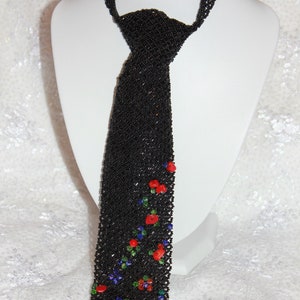 May include: A black beaded tie with a floral design. The tie is made of small black beads and features red, blue, and green accents. The tie is displayed on a white mannequin.