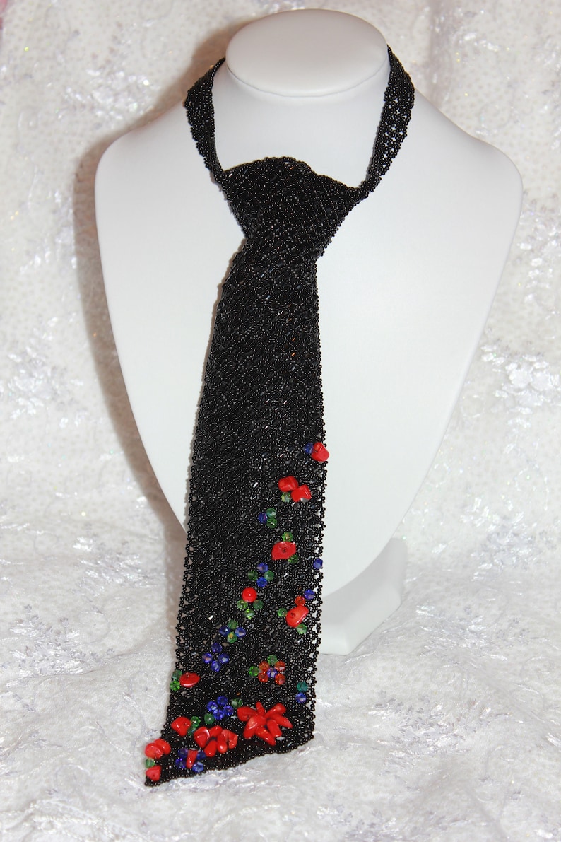 May include: A black beaded tie with a floral design. The tie is made of small black beads and features red, blue, and green accents. The tie is displayed on a white mannequin.