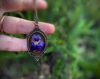 Op de afbeelding: Een delicate ketting met een ovale hanger van brons met een geperste paarse en gele bloem. De bloem is ingekapseld in helder hars en lijkt een viooltje te zijn.