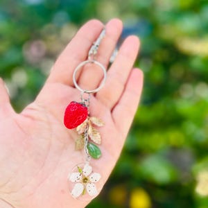Peut inclure: Un porte-cl&eacute;s argent&eacute; avec un charm en forme de fraise rouge, des feuilles dor&eacute;es, une perle verte et un pendentif circulaire transparent avec une fleur blanche. Le porte-cl&eacute;s est tenu dans une main sur un fond vert flou.