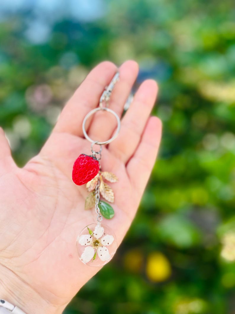 Peut inclure: Un porte-cl&eacute;s argent&eacute; avec un charm en forme de fraise rouge, des feuilles dor&eacute;es, une perle verte et un pendentif circulaire transparent avec une fleur blanche. Le porte-cl&eacute;s est tenu dans une main sur un fond vert flou.