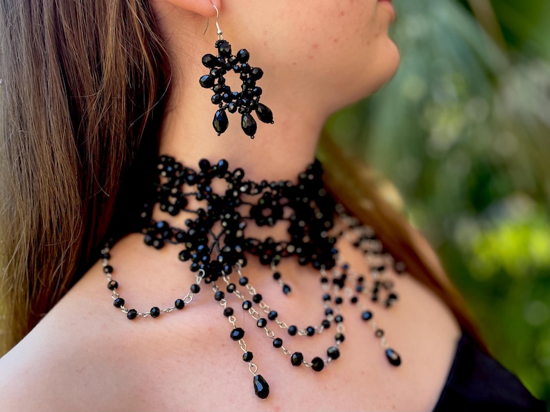 May include: A black beaded choker necklace with a delicate lace design and dangling strands of beads. The necklace is paired with matching black beaded earrings.