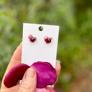 May include: A pair of heart-shaped earrings with pink and red pressed flower petals encased in clear resin.