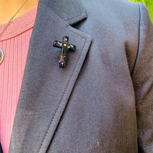 May include: A black blazer with a gold and black cross pin on the lapel.