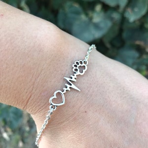 Puede incluir: Una pulsera de plata con un corazón, una huella de pata y un diseño de latido del corazón.
