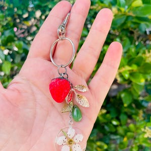 Peut inclure: Un porte-cl&eacute;s argent&eacute; avec un charm en forme de fraise rouge, des feuilles dor&eacute;es, des perles vertes et un pendentif circulaire transparent avec une fleur blanche. Le porte-cl&eacute;s est tenu dans une main sur un fond vert flou.