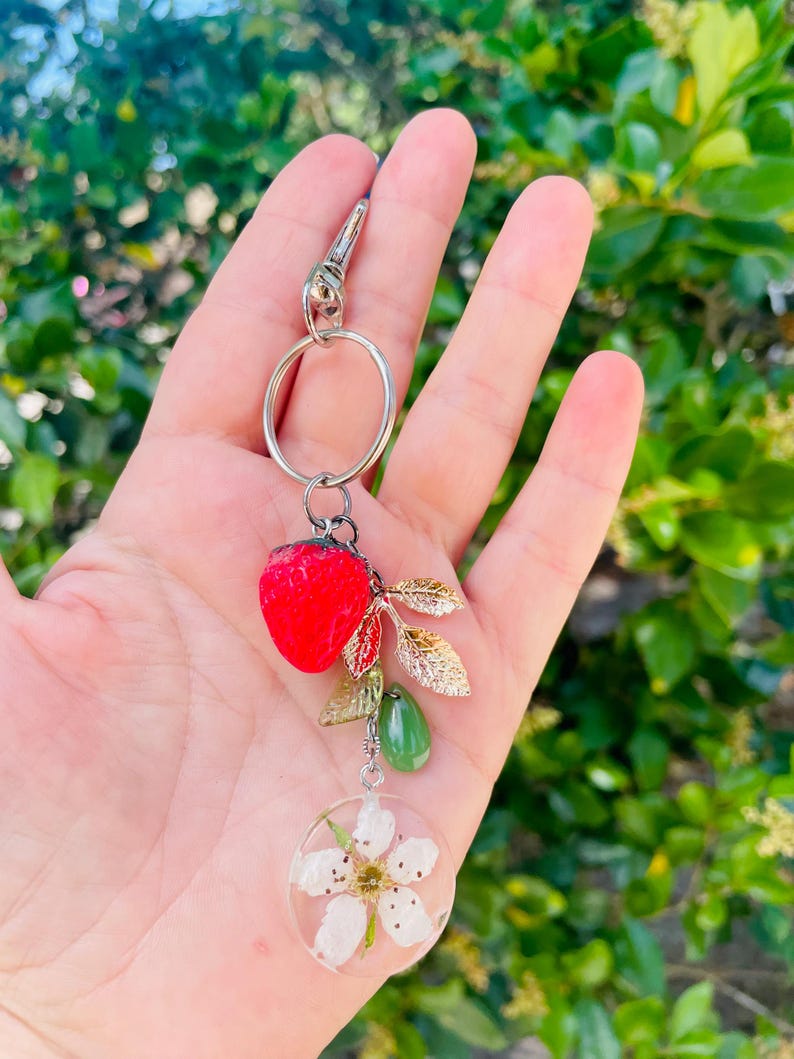 Peut inclure: Un porte-cl&eacute;s argent&eacute; avec un charm en forme de fraise rouge, des feuilles dor&eacute;es, des perles vertes et un pendentif circulaire transparent avec une fleur blanche. Le porte-cl&eacute;s est tenu dans une main sur un fond vert flou.