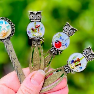 May include: A collection of decorative metal bookmarks. One bookmark features a starfish and seashells, while others showcase owl designs with miniature scenes of mushrooms, ladybugs, and flowers. The bookmarks have a vintage aesthetic.