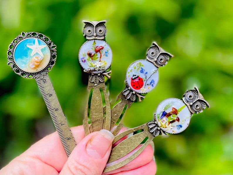 May include: A collection of decorative metal bookmarks. One bookmark features a starfish and seashells, while others showcase owl designs with miniature scenes of mushrooms, ladybugs, and flowers. The bookmarks have a vintage aesthetic.