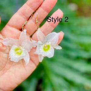 Pode incluir: Um par de brincos pendentes com p&eacute;talas brancas transl&uacute;cidas em forma de orqu&iacute;dea e um centro verde-lim&atilde;o vibrante. Os brincos est&atilde;o presos a ganchos de metal dourado. O texto "Style 2" &eacute; vis&iacute;vel no canto superior direito da imagem.