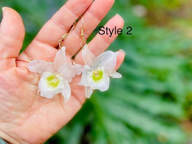 Pode incluir: Um par de brincos pendentes com p&eacute;talas brancas transl&uacute;cidas em forma de orqu&iacute;dea e um centro verde-lim&atilde;o vibrante. Os brincos est&atilde;o presos a ganchos de metal dourado. O texto "Style 2" &eacute; vis&iacute;vel no canto superior direito da imagem.