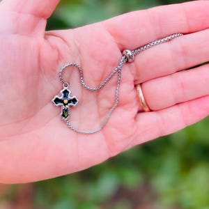 May include: A silver chain bracelet with a black cross charm. The cross has a gold center and green accents.