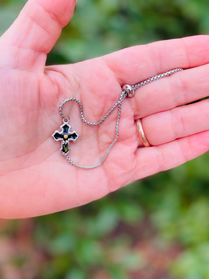 May include: A silver chain bracelet with a black cross charm. The cross has a gold center and green accents.