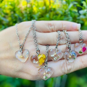 May include: Six silver necklaces with heart-shaped pendants filled with pressed flowers. The pendants are clear and show the flowers in detail. The necklaces are on a hand.